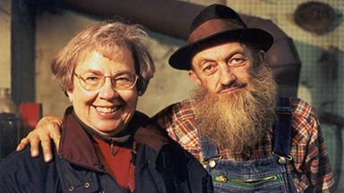 An old white man with a large beard poses with his arm around an elderly woman, both smiling at the camera