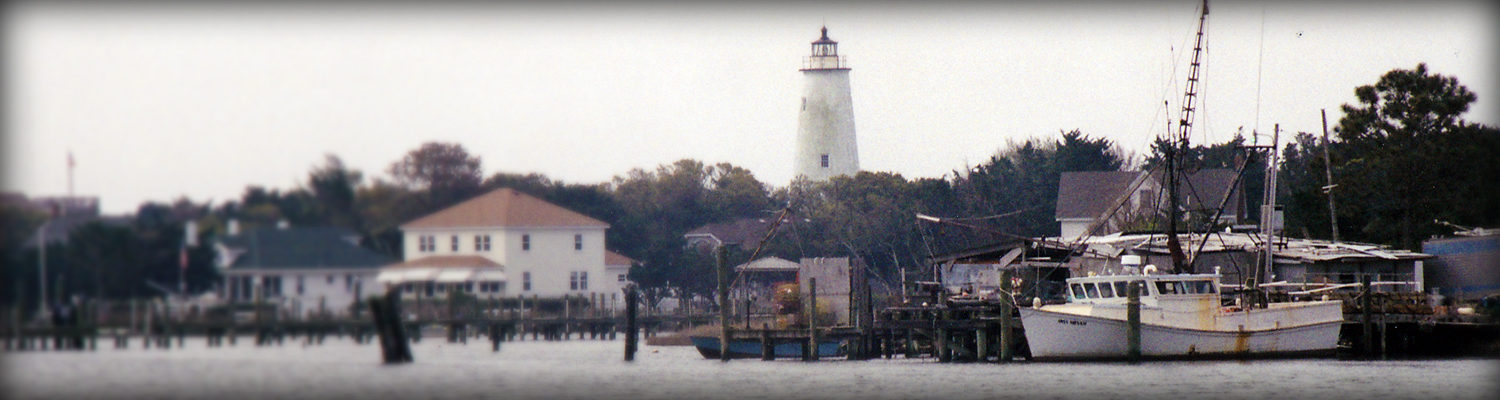 A somewhat rusted white fishing boat floats by a dock. Several more docks are seen next to it. Houses and trees are seen behind the harbor. In the background, a white lighthouse rises above the treeline.